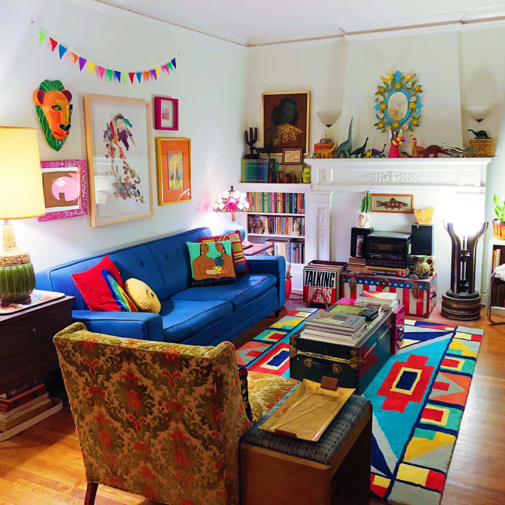 Colorful living room with blue sofa, vibrant rug, eclectic wall art, bookshelves, and plants around a white fireplace.