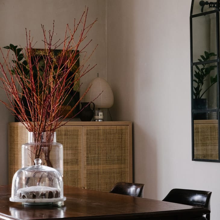 Dining room with a wooden table, glass vase with branches, wicker cabinet, and large grid mirror.
