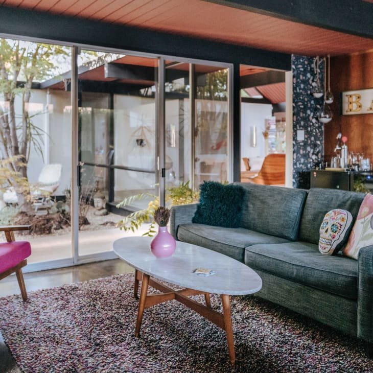 Mid-century modern living room with gray sofa, colorful pillows, pink chair, and sliding glass doors to a garden.