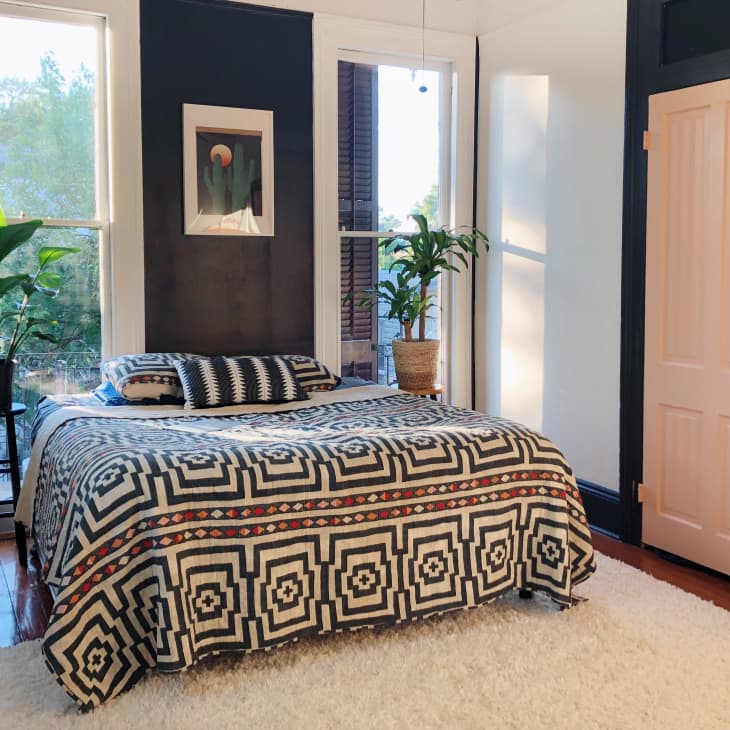 Bedroom with geometric patterned bedspread, two potted plants, and a framed artwork on dark walls.