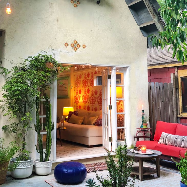 Cozy patio with red sofa, potted plants, and open French doors leading to a warmly lit living room with patterned wallpaper.