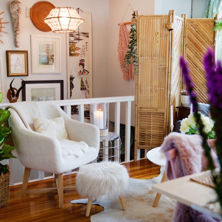 Cozy living room with white armchair, fluffy stool, potted plant, wall art, and bamboo room divider.