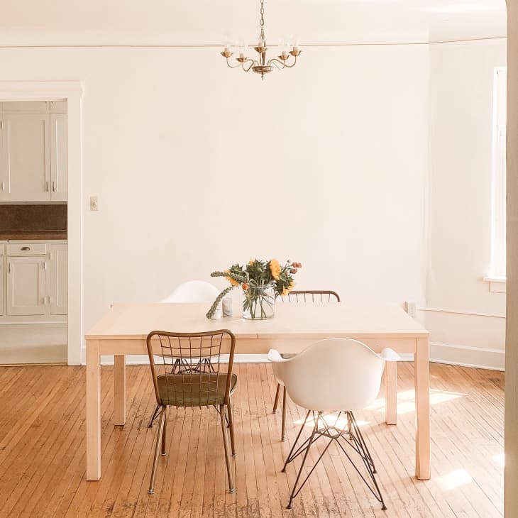 Dining room with light wood table, mixed chairs, floral centerpiece, and chandelier, adjacent to a kitchen with white cabinets.