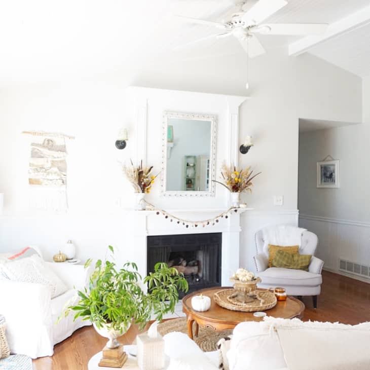 Bright living room with white sofas, plants, a fireplace, and a round mirror on the wall.