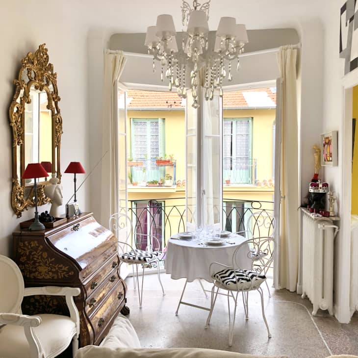 Elegant room with ornate mirror, chandelier, antique desk, and small dining table by a window with wrought iron chairs.