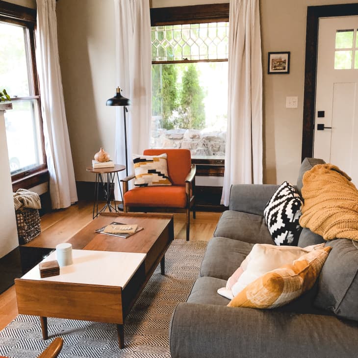 Cozy living room with gray sofa, orange armchair, wooden coffee table, and large windows with white curtains.