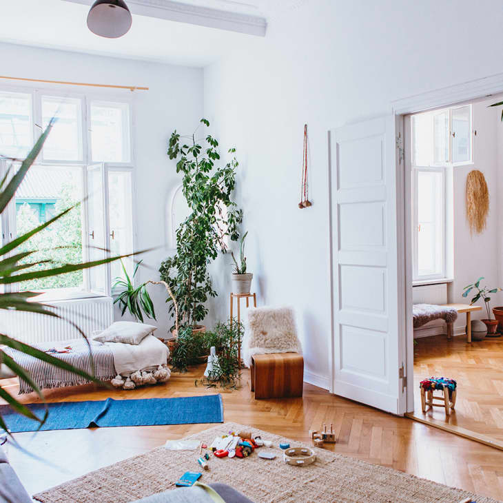 Bright living room with large plants, wooden floors, and cozy seating, leading to a sunlit room with more greenery.