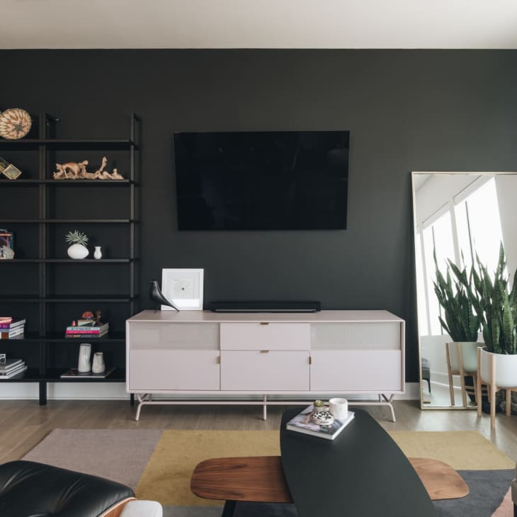 Living room with black wall, TV, white console, large mirror, plant, and black shelving with decor items.