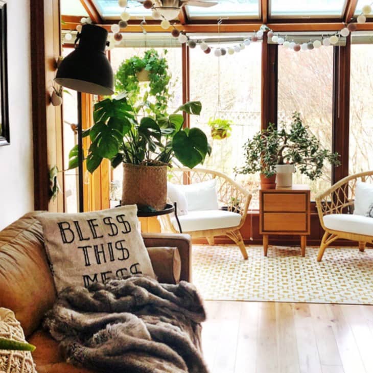 Sunroom with rattan chairs, large plants, a wooden side table, and a cozy sofa with a "Bless This Mess" pillow.
