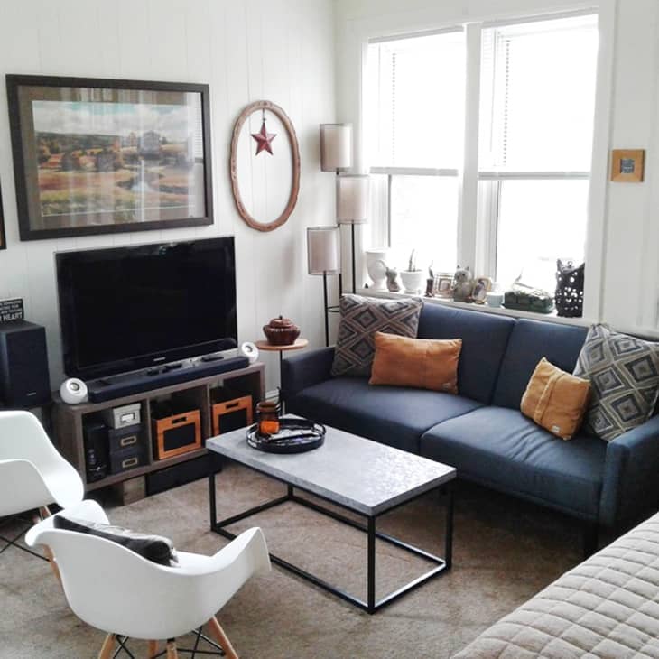 Living room with blue sofa, orange cushions, white chairs, TV, and wall art, featuring a modern coffee table and decor items.