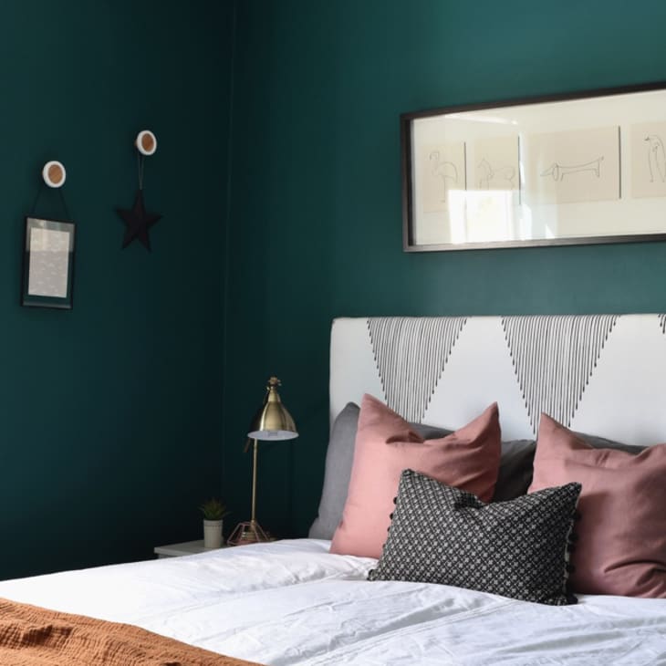 Teal bedroom with white headboard, pink and gray pillows, gold lamp, and framed art on the wall.