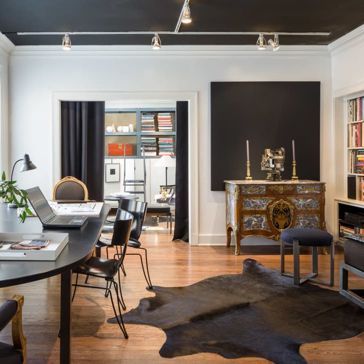 Home office with black ceiling, bookshelves, ornate dresser, black cowhide rug, and a round table with laptops and decor.