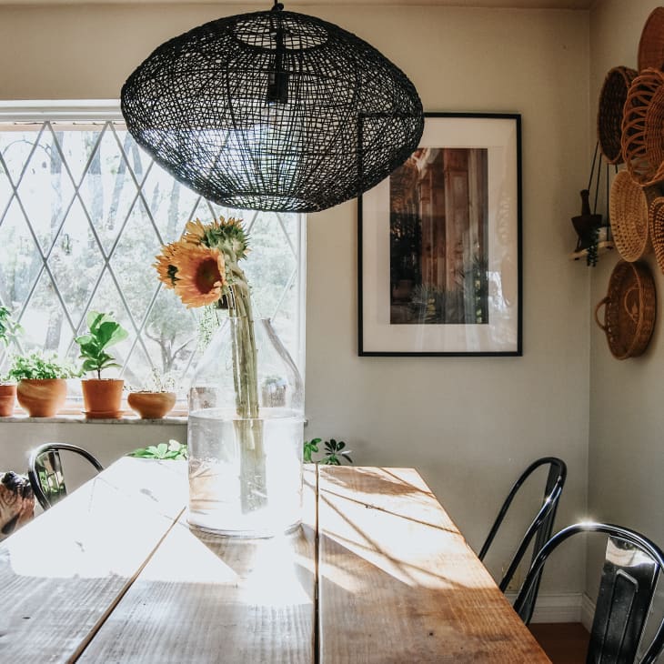 Dining room with wooden table, black chairs, sunflowers in vase, wicker baskets on wall, and potted plants by window.