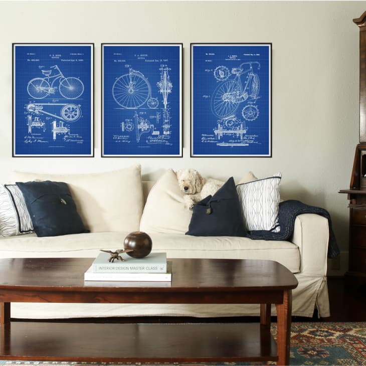 Living room with cream sofa, blue pillows, wooden coffee table, and three bicycle blueprint posters on the wall.