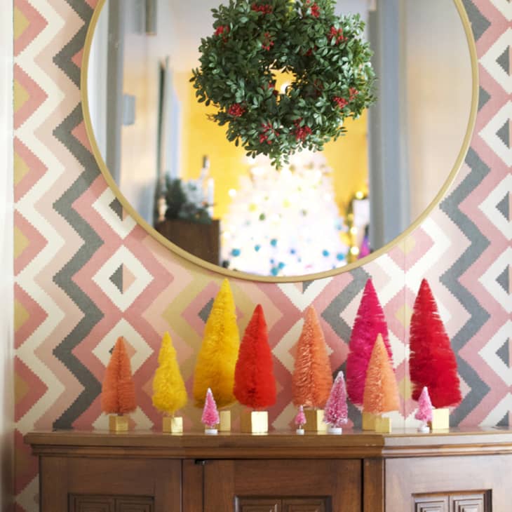 Colorful mini Christmas trees on a wooden cabinet, with a round mirror and wreath above, against geometric wallpaper.