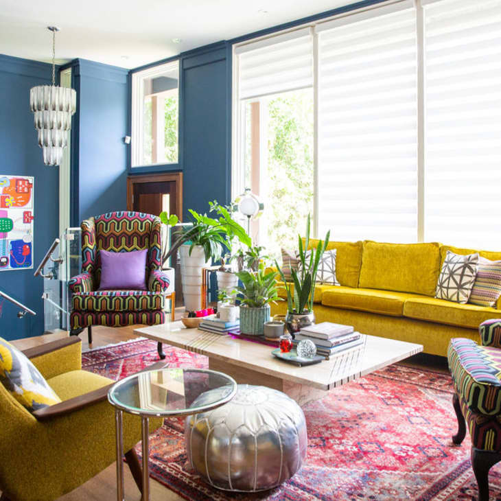 Colorful living room with yellow sofa, patterned armchairs, blue walls, and a red rug, featuring plants and modern decor.
