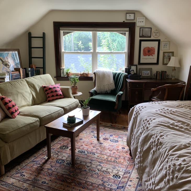 Cozy attic bedroom with a green sofa, patterned rug, wooden desk, and plants by a window.