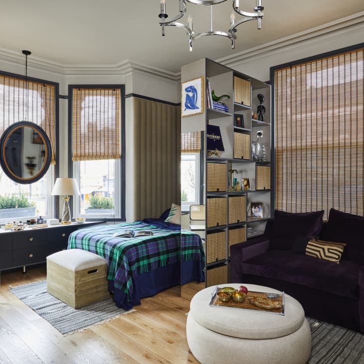 An apartment with bamboo blinds and a tall bookshelf with wicker baskets and open shelving separating the living space from the bed