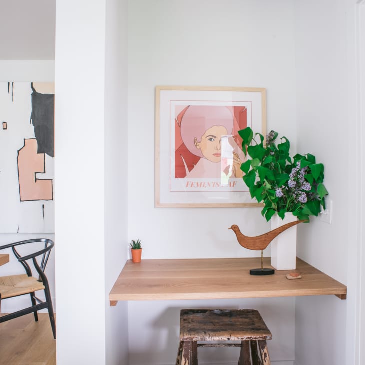 Small wooden desk with a plant, bird sculpture, and framed art in a modern room with abstract wall art and dining chairs.