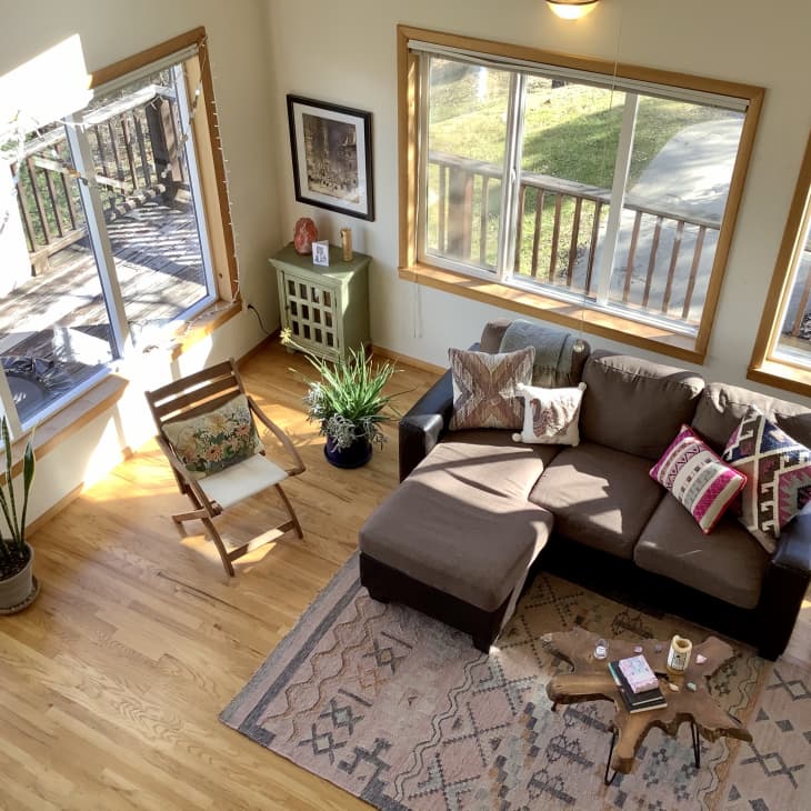 Cozy living room with brown sectional sofa, patterned pillows, wooden chair, plants, and a rustic coffee table on a patterned rug.