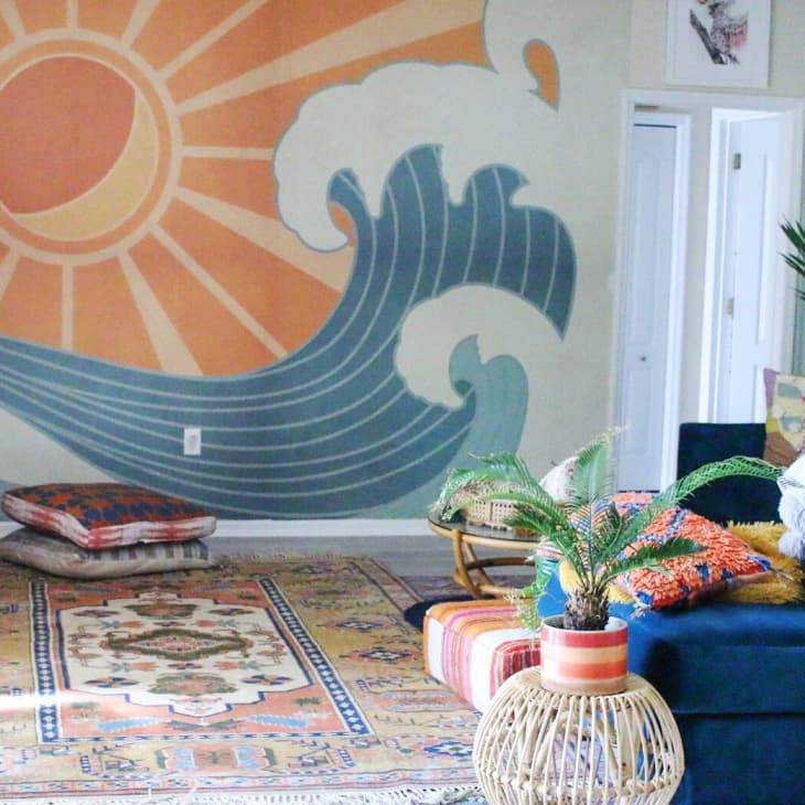 Living room with sun and wave mural, colorful cushions, blue sofa, and potted plants on a patterned rug.