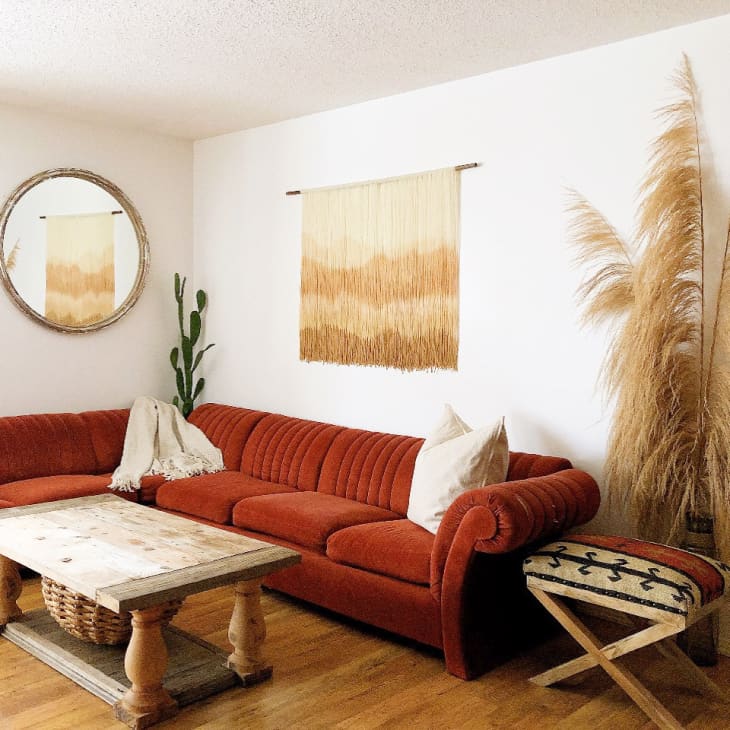 Rust-colored sectional sofa in living room with wooden coffee table, round mirror, wall hanging, and pampas grass decor.