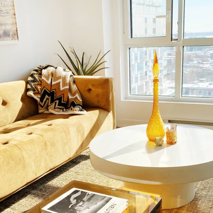 Sunny living room with a mustard sofa, chevron throw, round white table, and tall amber vase near a large window.