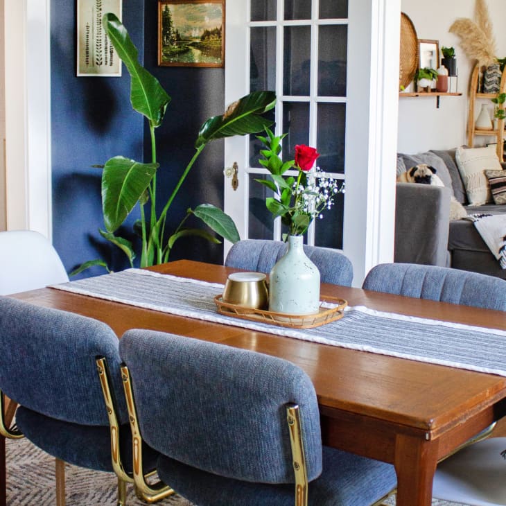 Dining room with wooden table, blue chairs, vase with red rose, and view into living room with gray sofa and plants.