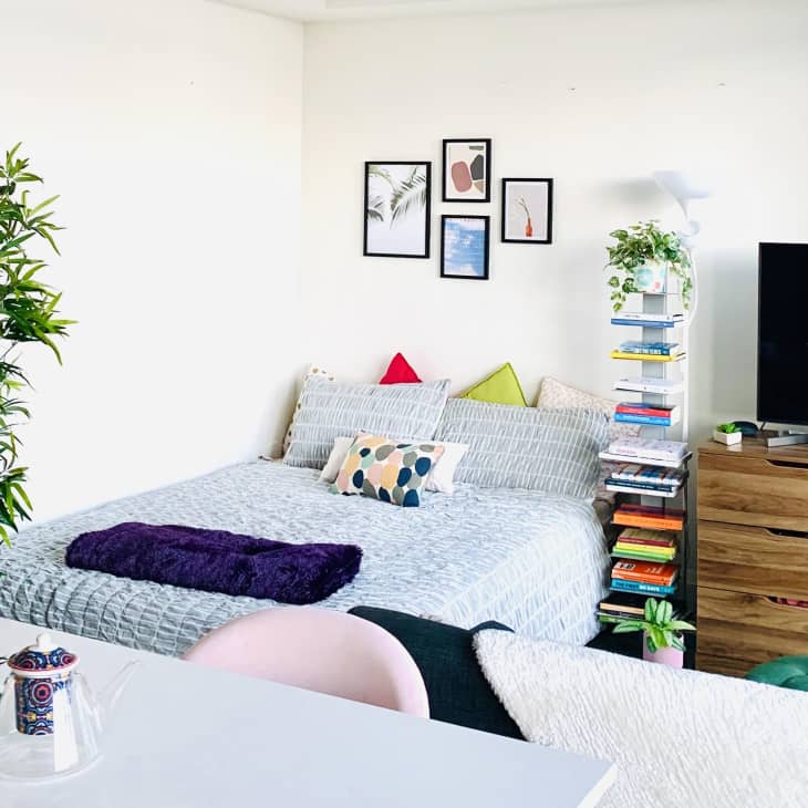Bedroom with a bed, colorful pillows, wall art, TV on a wooden dresser, and a plant. Books stacked on a shelf.