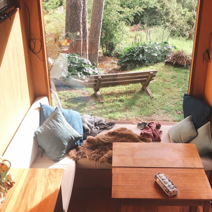 Cozy sunlit nook with a wooden table, cushions, and a view of a garden with a bench.