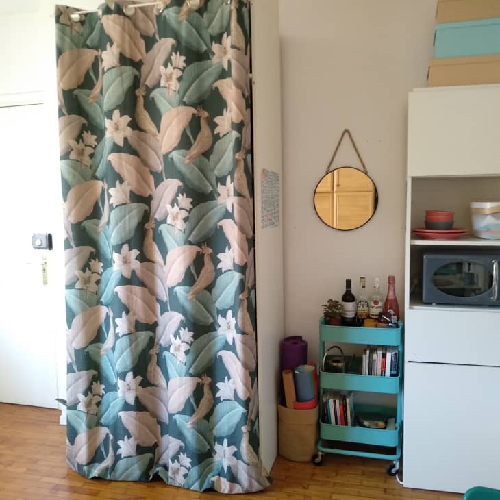 Floral curtain, round mirror, microwave on shelf, teal cart with bottles and books, and stacked boxes in a small room.