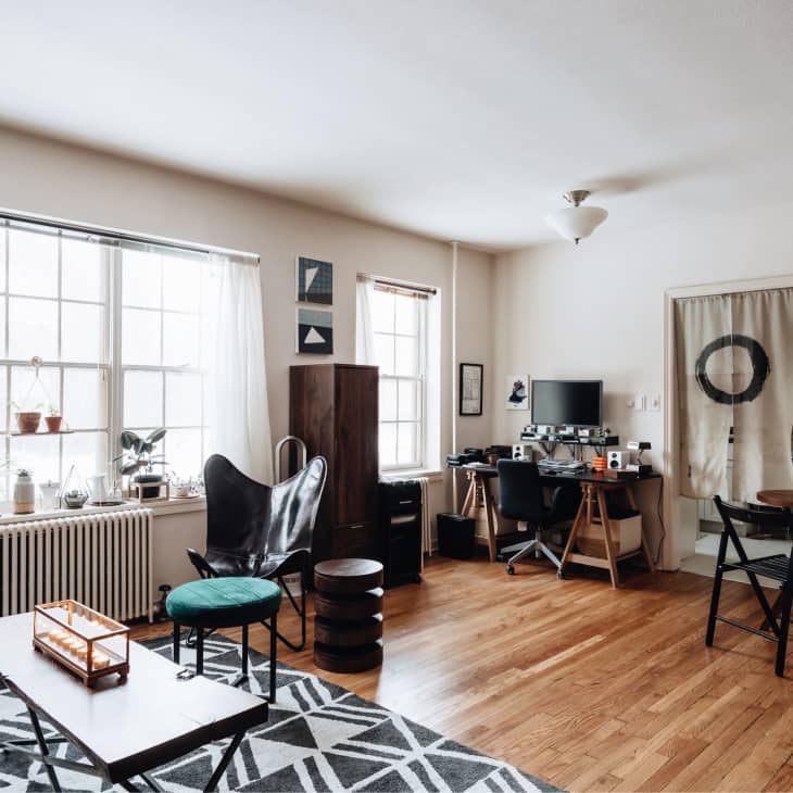 Living room with wooden floors, black chair, desk with computer, round dining table, and large windows with plants.