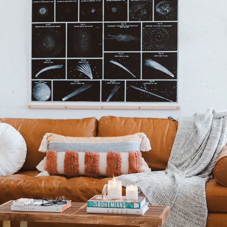 Tan leather sofa with textured pillows and a gray throw, under a space-themed wall art. Coffee table with books and candles.