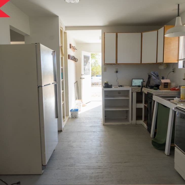 Outdated kitchen with white cabinets, fridge, stove, and cluttered countertops under a window with blinds.
