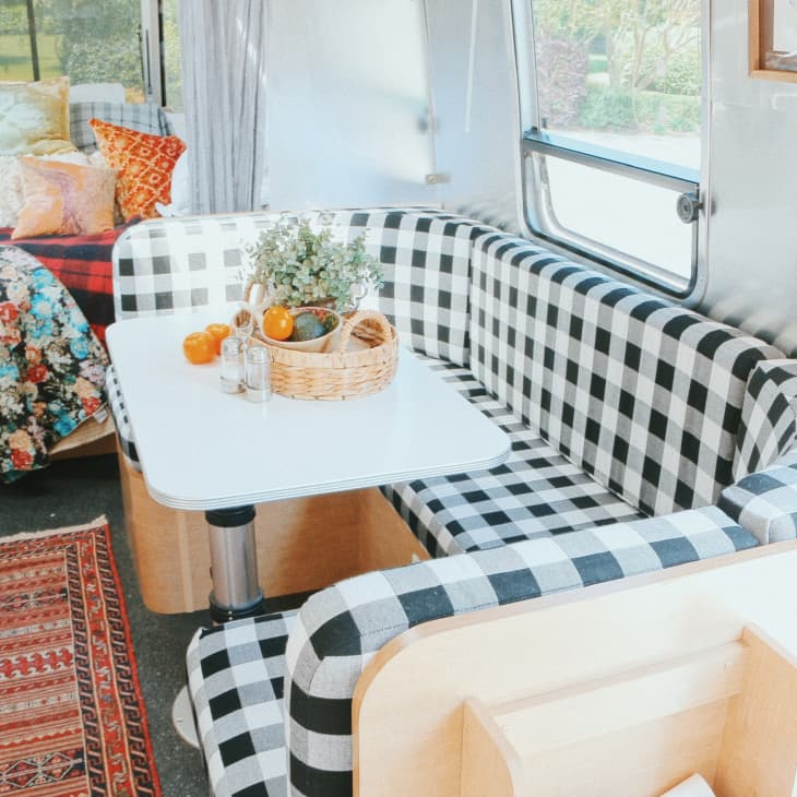 Camper interior with black and white checkered seating, white table, basket of fruit, and colorful bedding.