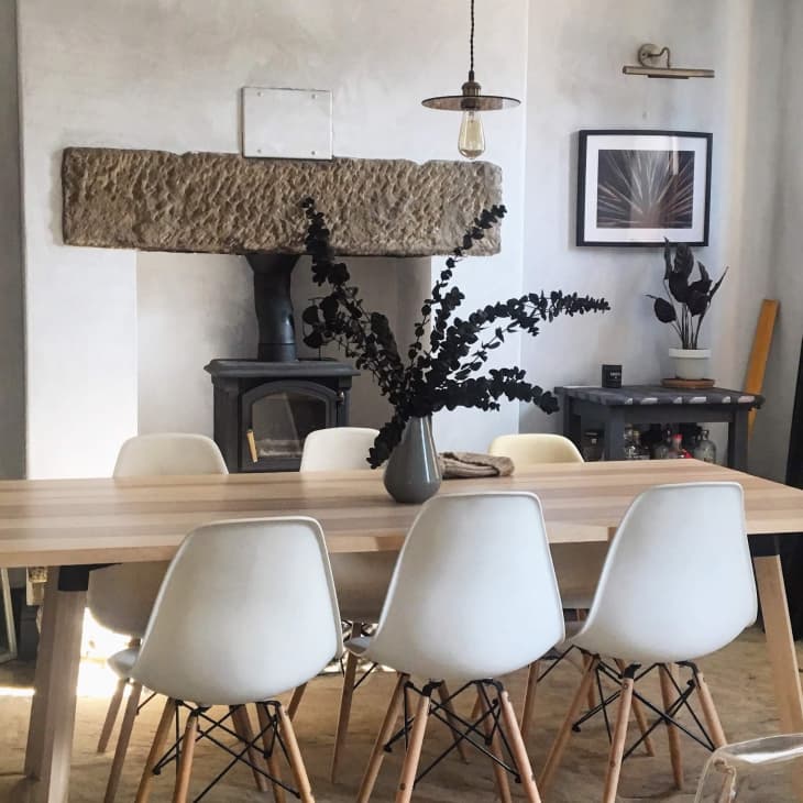 Dining room with a wooden table, six white chairs, a vase with dark branches, and a wood-burning stove under a stone mantel.