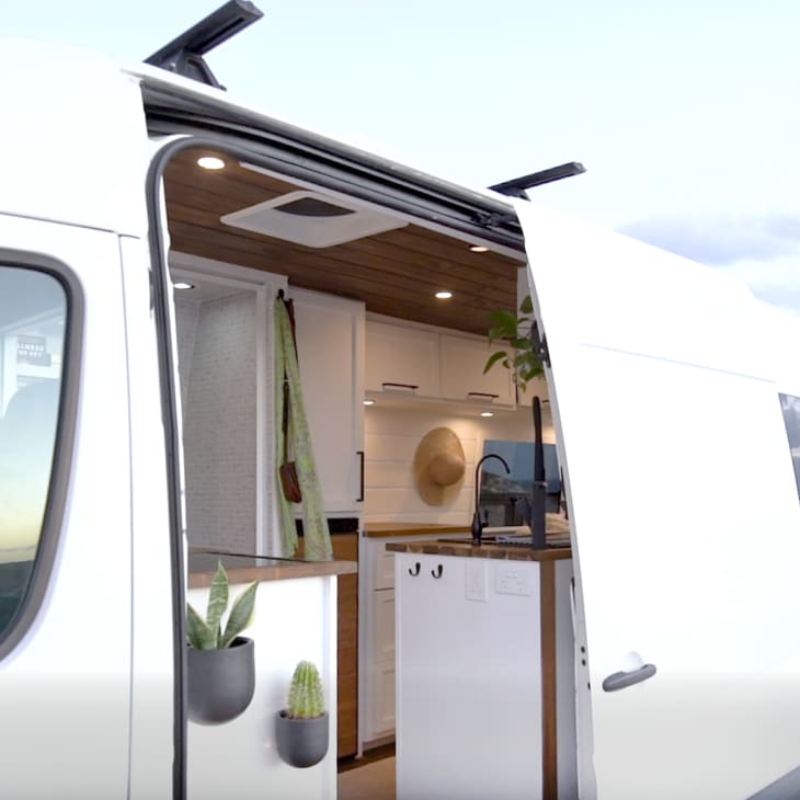 Camper van with open sliding door, showing interior kitchen, plants, and ocean view in the background.