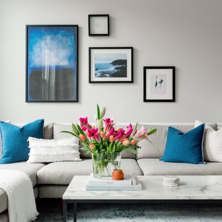 Living room with a beige sectional sofa, blue pillows, marble coffee table, and vibrant pink flowers. Art frames on the wall.