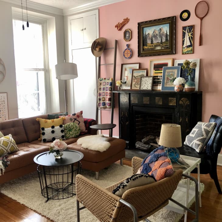 Cozy living room with a brown sofa, colorful pillows, pink accent wall, and eclectic decor above a black fireplace.