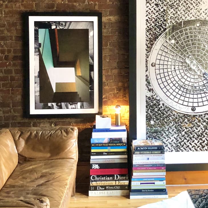 Brown leather sofa, brick wall with abstract art, stack of books, and a small lamp on a wooden floor.