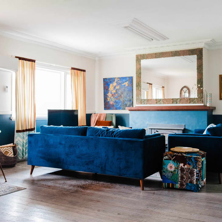 Living room with blue velvet sofas, large mirror, colorful paintings, and a wooden dining table with chairs.