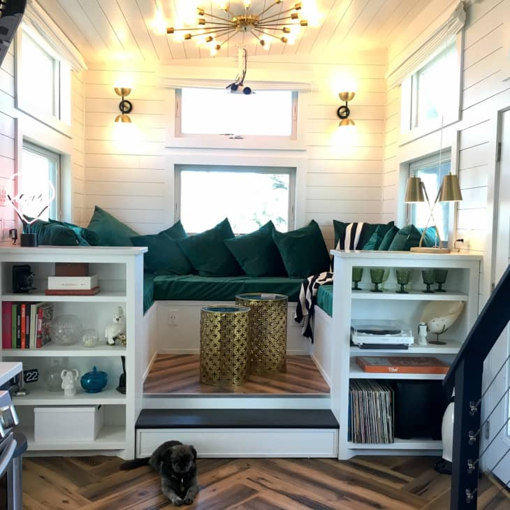 Cozy tiny house living area with green cushions, gold tables, bookshelves, and a cat on herringbone wood floor.