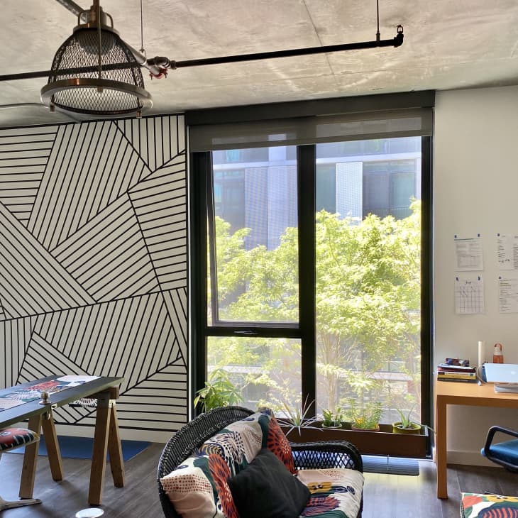 Modern office with geometric wall art, large windows, blue chair, wooden desk, and colorful cushions on a black sofa.
