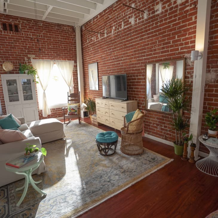 Loft living room with exposed brick walls, beige sectional sofa, TV on dresser, plants, and natural light from large windows.