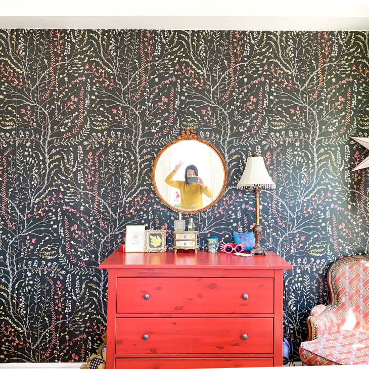 Red dresser with a round mirror, floral wallpaper, table lamp, and patterned armchair in a bright room.