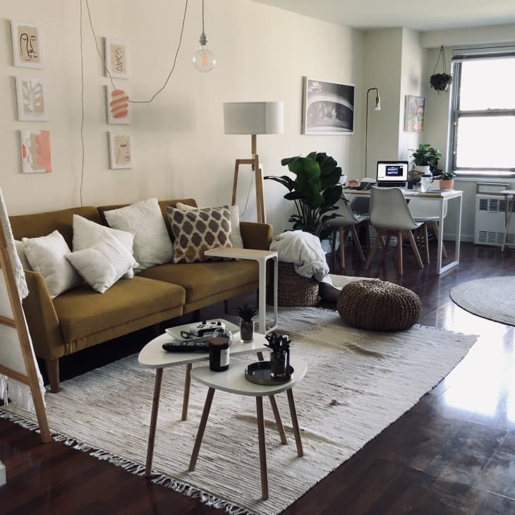 Living room with mustard sofa, white pillows, ladder shelf, round tables, and a workspace by the window with a laptop.