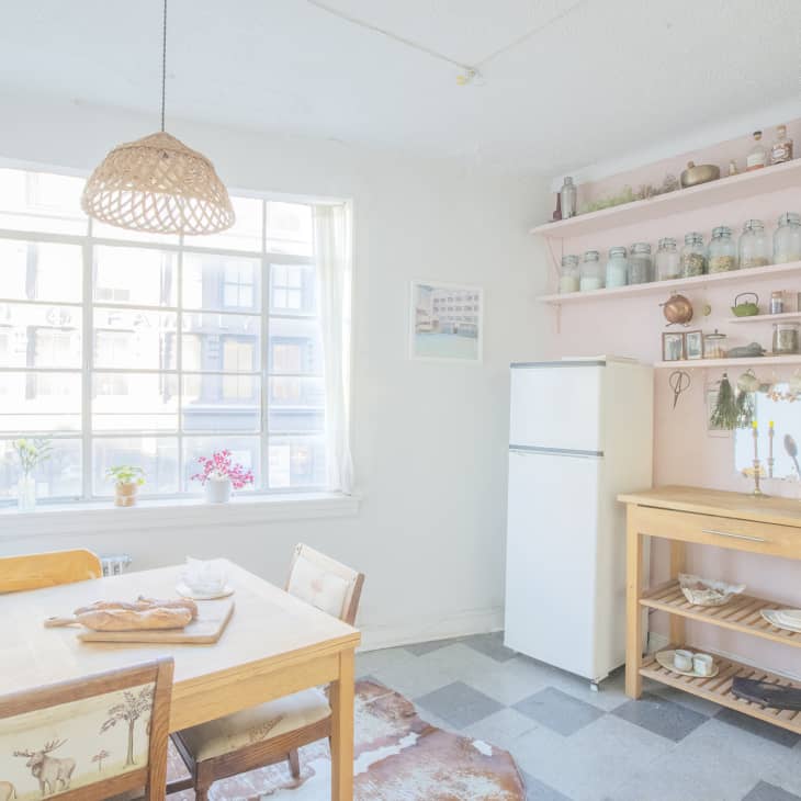 Bright kitchen with wooden dining table, bread on a cutting board, potted plants on windowsill, and pink shelves with jars.