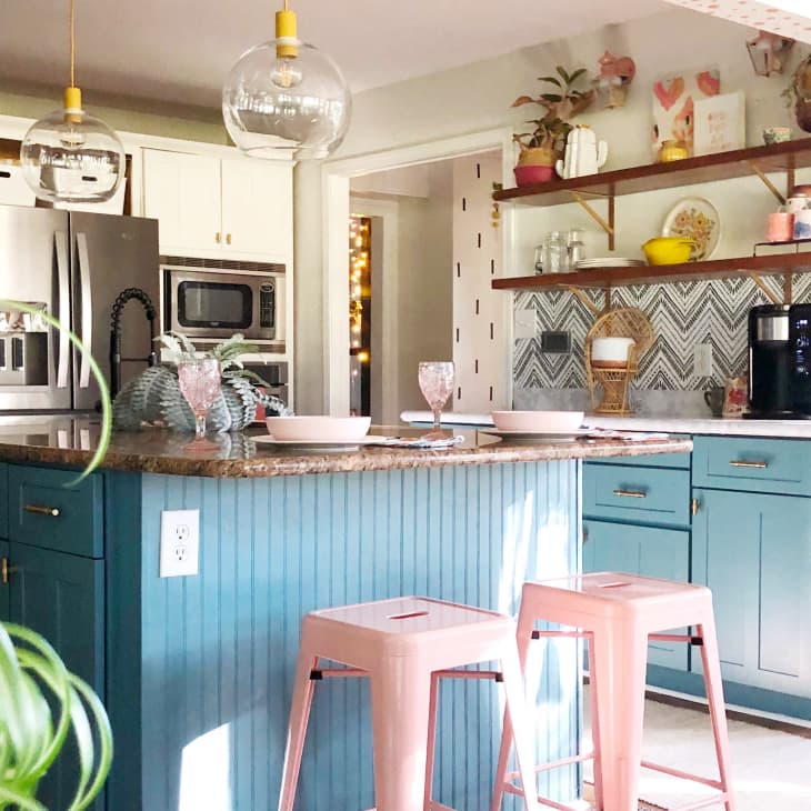 Bright kitchen with teal cabinets, pink stools, granite island, and open shelves displaying decor and plants.