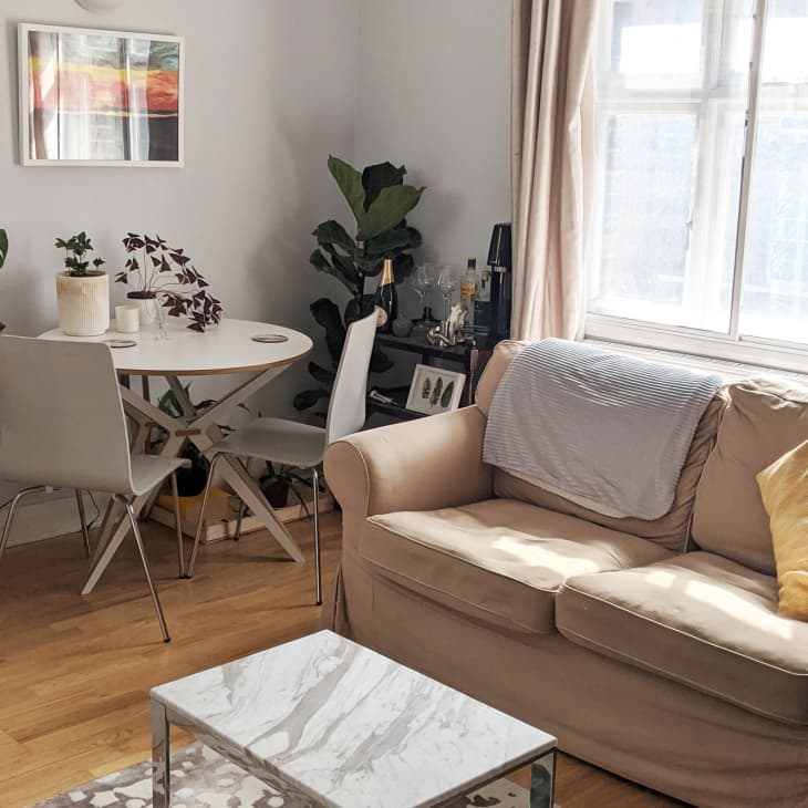 Cozy living room with beige sofa, yellow pillow, small round dining table, white chairs, and potted plants by the window.