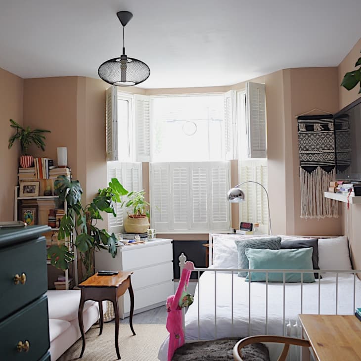 Cozy bedroom with plants, bookshelves, a white bed, wall art, and a TV. Light filters through bay windows with shutters.